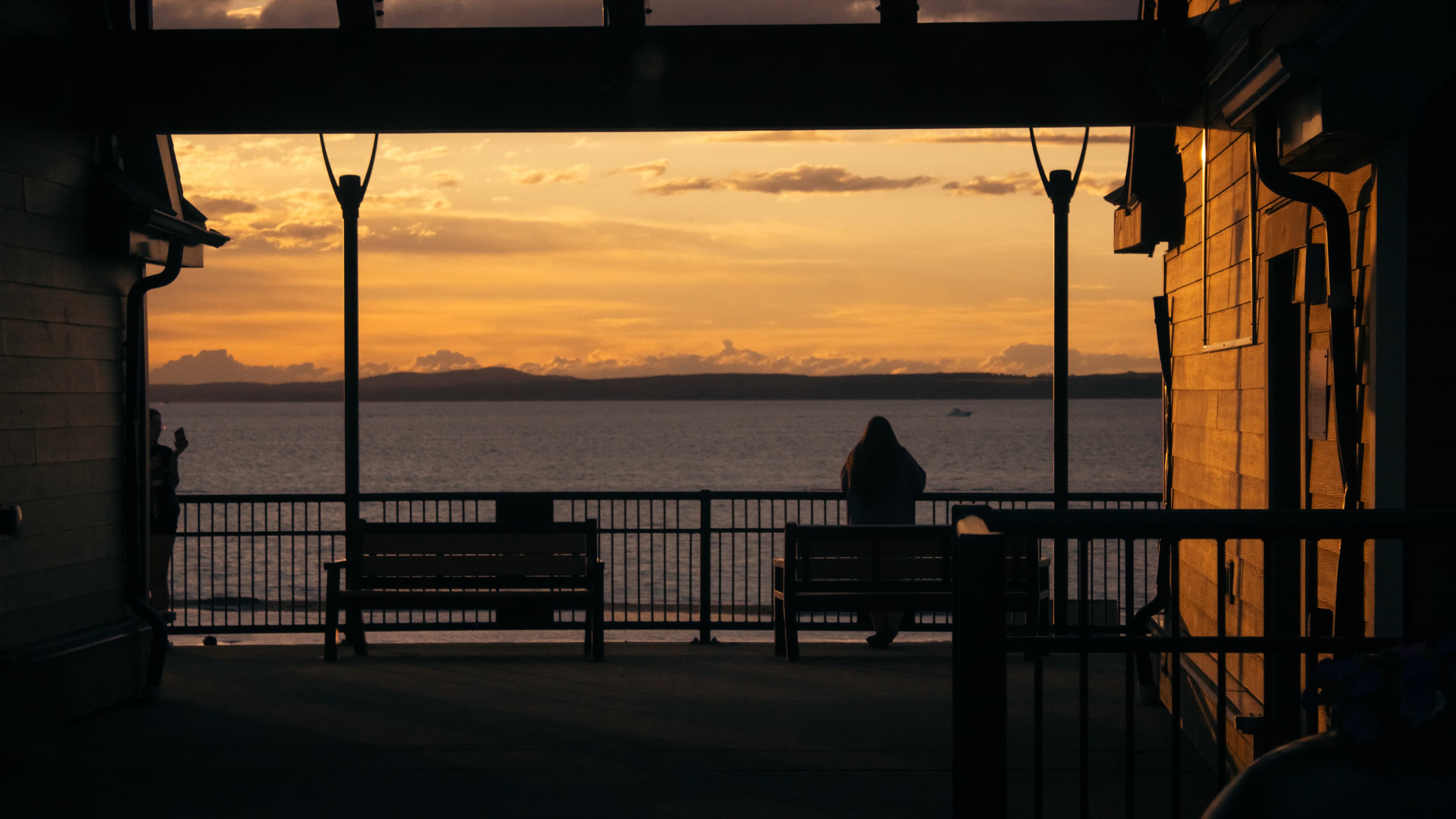 Golden hour over the water — a lone figure at the railing, framed by a waterfront pavilion